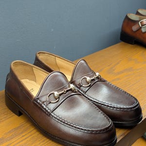 May include: A pair of brown leather loafers with silver-colored metal accents. The shoes have a classic design with a rounded toe and a decorative strap across the top. The loafers are displayed on a wooden surface.