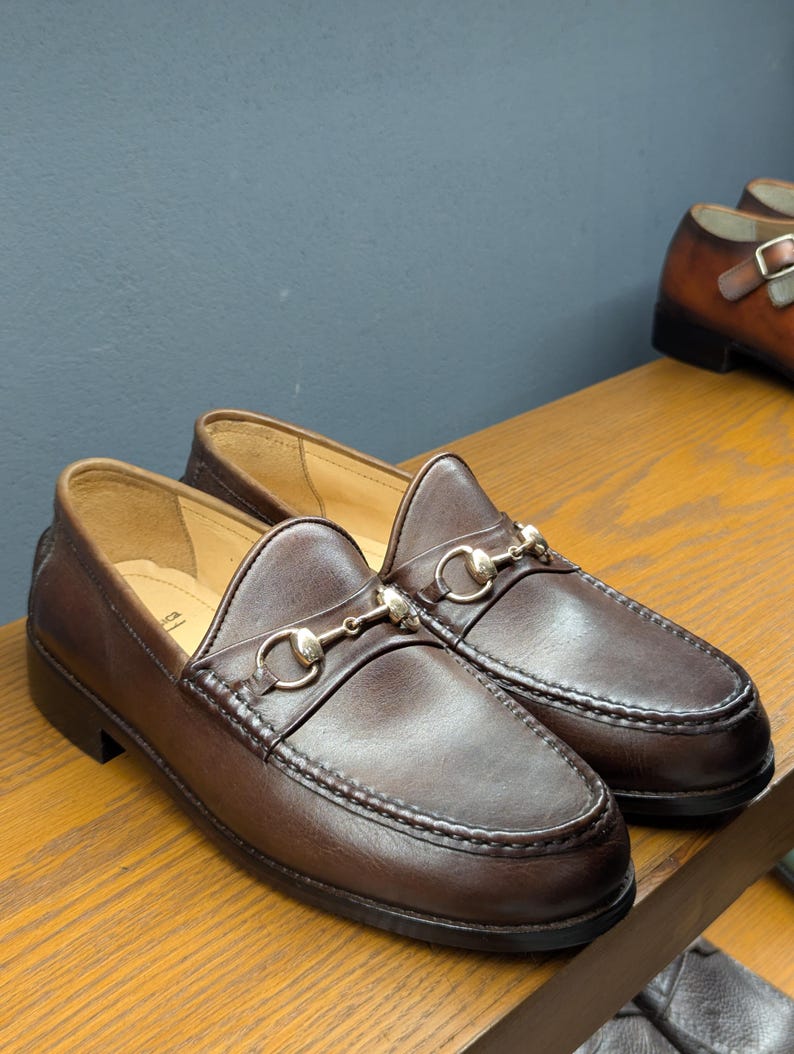 May include: A pair of brown leather loafers with silver-colored metal accents. The shoes have a classic design with a rounded toe and a decorative strap across the top. The loafers are displayed on a wooden surface.