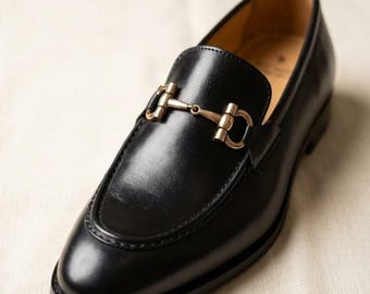 May include: A single black leather loafer with a gold-colored metal buckle. The shoe has a rounded toe and a low heel. The loafer is displayed on a neutral-colored surface.