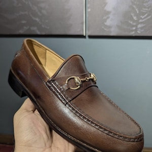 May include: Brown leather loafer with a gold-tone metal horsebit detail. The shoe features a rounded toe, a slip-on design, and a stitched sole. The leather has a polished finish, and the shoe is held by a hand.