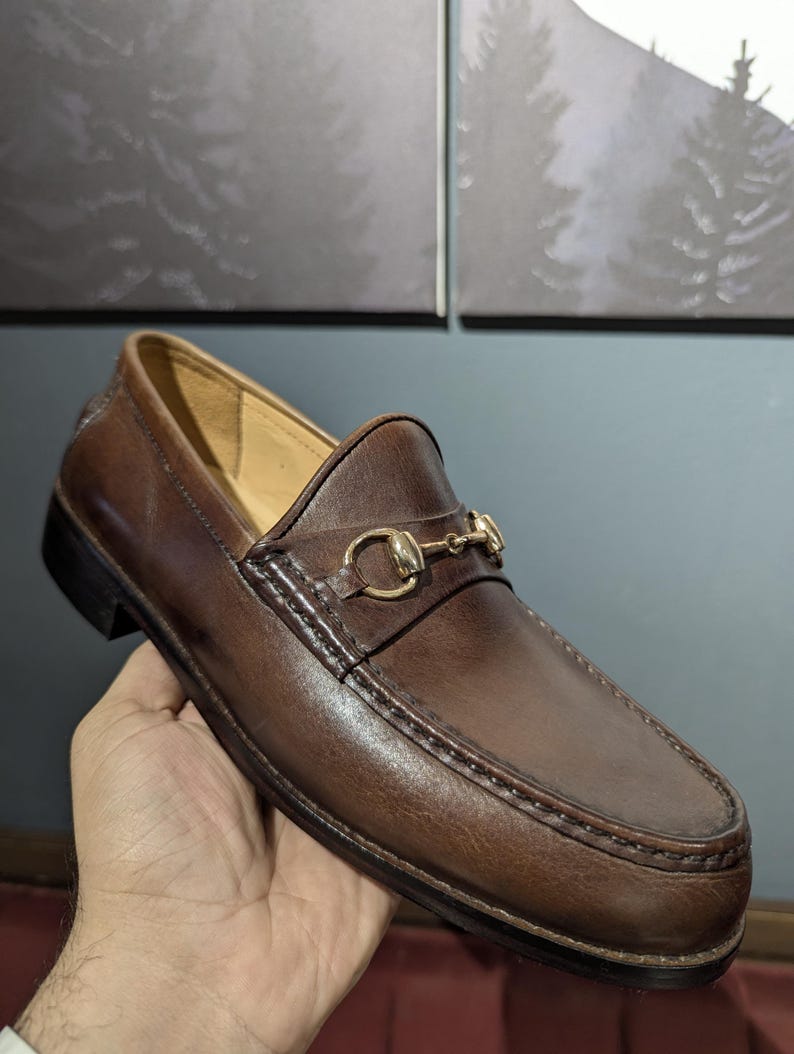 May include: Brown leather loafer with a gold-tone metal horsebit detail. The shoe features a rounded toe, a slip-on design, and a stitched sole. The leather has a polished finish, and the shoe is held by a hand.