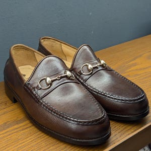 May include: A pair of brown leather loafers with a classic design. Each shoe features a decorative metal bit across the vamp. The shoes are displayed on a wooden surface against a dark background.