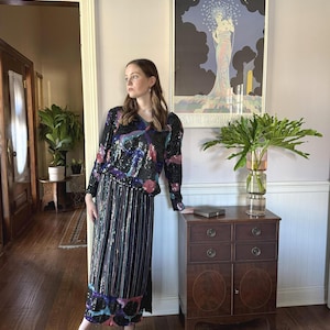 May include: A woman wearing a black sequined two-piece outfit with a long-sleeved top and a floor-length skirt. The outfit features colorful sequin accents. She is wearing black heels. A vintage poster hangs on the wall behind her.