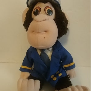 May include: A plush monkey toy wearing a blue pilot uniform jacket with yellow stripes and a white captain hat featuring a crest emblem. The monkey has dark brown fur, light beige skin, and large eyes with orange and black circular pupils.