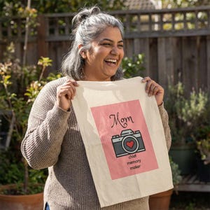 Può includere: Un canovaccio color crema con un design rettangolare rosa con una grafica di fotocamera e le parole "Mamma" e "chief memory maker". La fotocamera ha un cuore rosso al centro. L'asciugamano è tenuto da una persona.