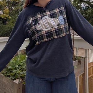 May include: A dark charcoal long-sleeve henley shirt featuring a rectangular patch on the chest. The patch is made of green, navy, and white plaid fabric with a stitched applique of a fish in beige and blue fabric with a black eye detail.