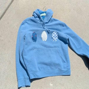 Op de afbeelding: Een lichtblauwe hoodie met een kangoeroezak en een capuchon met trekkoord. De hoodie heeft drie surfboard-vormige patches in denim, witte gewatteerde stof en een blauw-wit geblokt patroon. De hoodie hangt aan een hanger.