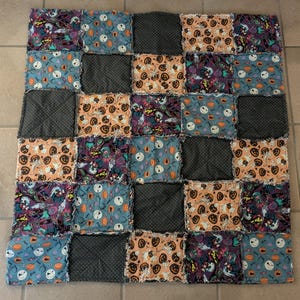 May include: A patchwork quilt featuring a Halloween theme. The quilt is composed of squares in various colors and patterns, including orange pumpkins, purple bats, and teal ghosts. The squares are arranged in a grid pattern, with dark gray squares interspersed throughout.