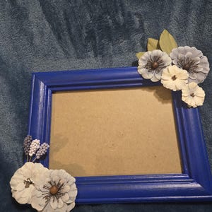 May include: A rectangular blue picture frame decorated with paper flowers in shades of white, gray, and purple. The frame has a painted finish and a neutral-colored backing.