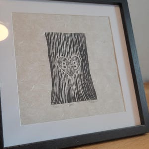 Op de afbeelding: Zwart-wit ingelijste print met een boomstamontwerp met een hartvormige gravure met de letters "B+B". De print staat tegen een getextureerde, gebroken witte achtergrond en zit in een zwarte lijst.