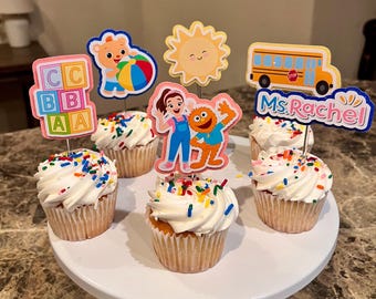 Könnte beinhalten: Cupcakes mit weißem Zuckerguss und bunten Streuseln, belegt mit dekorativen Ausschnitten eines Bären, einer Sonne, eines Schulbusses, Buchstabenblöcken und Figuren. Die Cupcakes sind auf einem weißen Kuchenständer angeordnet.