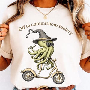 Può includere: Una t-shirt color crema con la grafica di un polpo verde chiaro che indossa un cappello da strega grigio scuro e occhiali da sole neri mentre guida un monopattino giallo. Il testo "Off to commithom foolery" è stampato in marrone sopra.