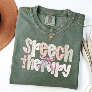 Puede incluir: Una camiseta verde oliva con las palabras "Speech therapy" en un diseño de patchwork con telas florales y estampadas. Un lazo rosa está centrado debajo de la palabra "Speech". Un collar dorado con un colgante está drapeado sobre la camisa.