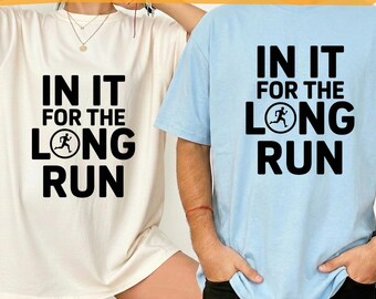 Puede incluir: Dos camisetas, una crema y otra azul claro, cada una con el texto "IN IT FOR THE LONG RUN" en negro. El diseño incluye una figura corriendo dentro de un círculo. Las camisetas son probablemente para una pareja o un equipo.