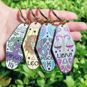 May include: Four colorful keychains featuring zodiac signs: Virgo, Leo, Pisces, and Libra. Each keychain has a gold-colored ring and a unique design with astrological symbols and names. The keychains are held in a hand against a green background.