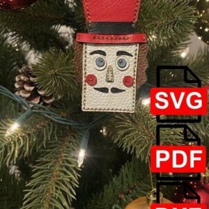 Puede incluir: Adorno navideño de cuero con forma de cascanueces, con sombrero de copa rojo y banda negra. El adorno tiene una cara color crema con ojos azules, bigote negro y mejillas rojas. El adorno está colgado de un árbol de Navidad.