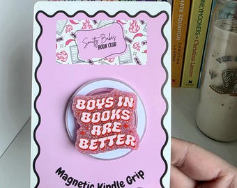 May include: A pink magnetic Kindle grip on a scalloped backing card. The grip features a glittery pink surface with the red text "BOYS IN BOOKS ARE BETTER". The card includes a "Smutty Babes BOOK CLUB" logo and the text "Magnetic Kindle Grip".