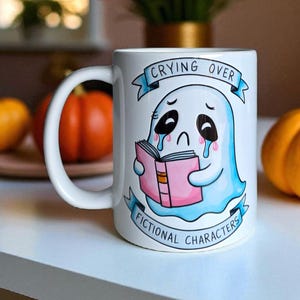 May include: White ceramic mug with a cartoon ghost design. The ghost is crying and holding a pink book. The text on the mug reads "CRYING OVER FICTIONAL CHARACTERS" in blue banners. The mug has a white handle.