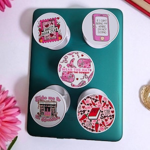 May include: Five white and pink phone grips with different designs and text. The designs include a book club theme, a phone with text, cowboy boots, a bookstore, and chili peppers. The grips are displayed on a teal surface.