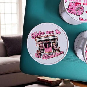 May include: Round white pop sockets with pink and white graphics. One says "Take me to the Bookstore" with a bookstore illustration. Another says "Wear the Ride the" with a hat and boot design. The pop sockets are attached to a teal surface.