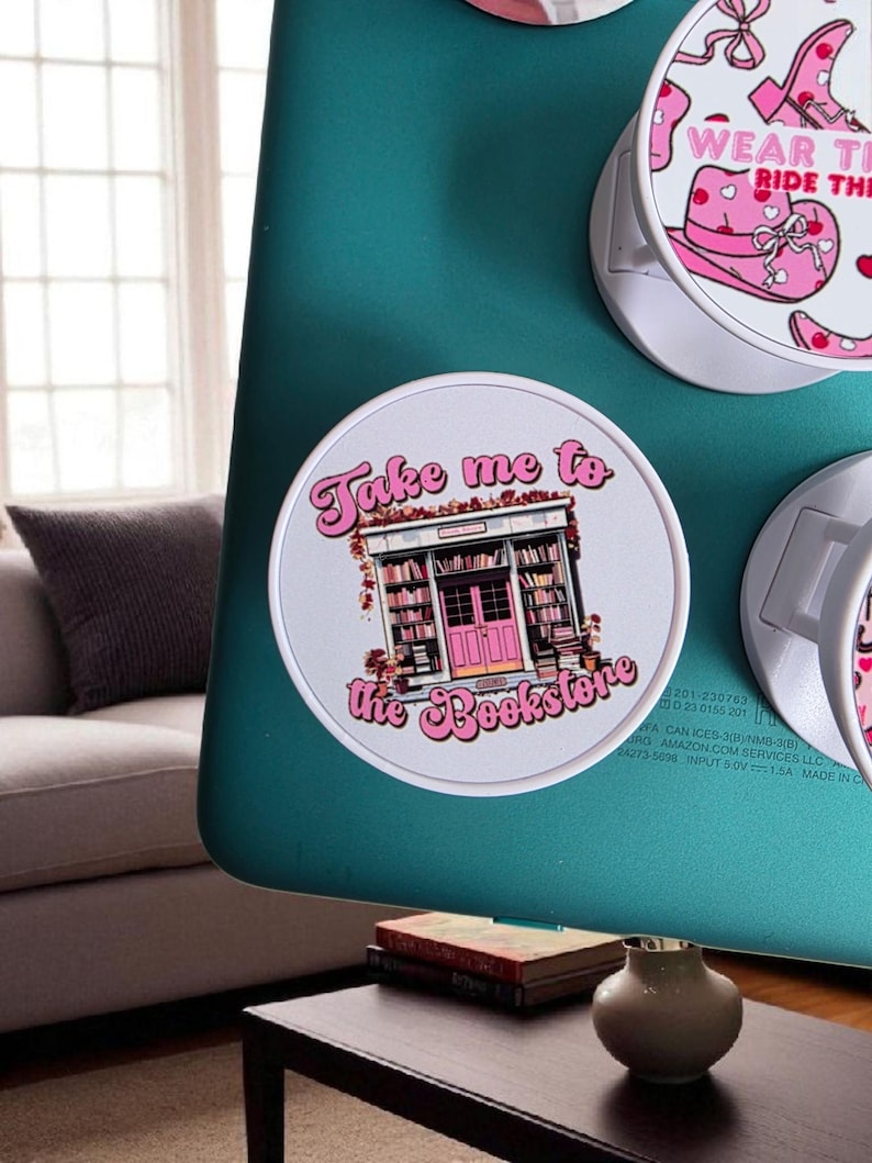 May include: Round white pop sockets with pink and white graphics. One says "Take me to the Bookstore" with a bookstore illustration. Another says "Wear the Ride the" with a hat and boot design. The pop sockets are attached to a teal surface.