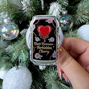 May include: A clear acrylic pin shaped like a can, featuring a black background with a red heart design and the text "Dark Romance - Forbidden Cherry". The pin is decorated with roses and chains, held in front of a Christmas tree.