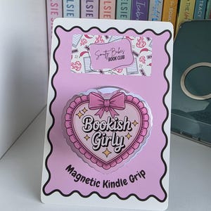 May include: A pink magnetic Kindle grip with a heart design. The grip has the text "Bookish Girly" and "Magnetic Kindle Grip". The top of the card has the text "Smutty Babes Book Club".