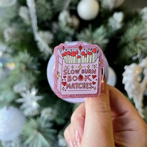 May include: A pink and glittery book of matches with the text "SLOW BURN BOOK MATCHES". The match heads are red, and the box has a pink glitter border. The background is a blurred Christmas tree with white baubles.