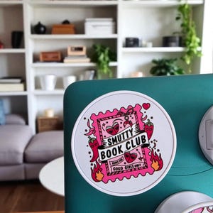 May include: A round white sticker with a pink and red design. The sticker features the text "Smutty Book Club" and "Good Girls Only". The design includes hearts, chili peppers, and flames.