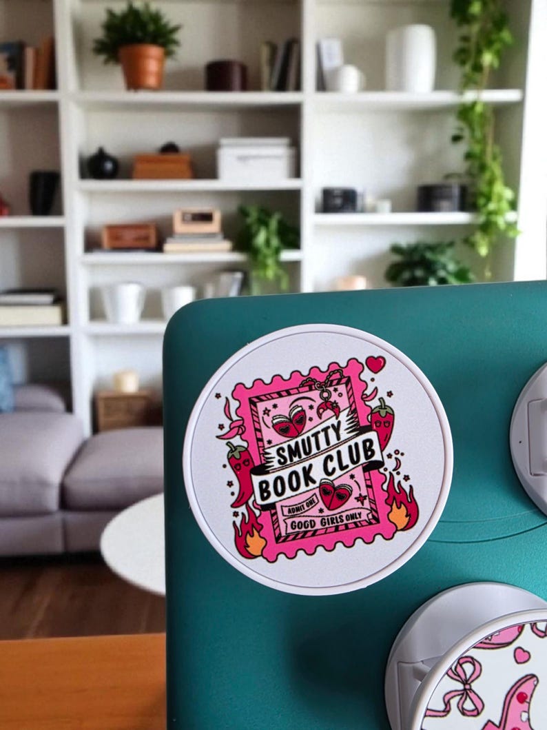 May include: A round white sticker with a pink and red design. The sticker features the text "Smutty Book Club" and "Good Girls Only". The design includes hearts, chili peppers, and flames.