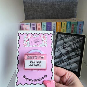 May include: A pink magnetic Kindle grip with the text "HELLO I'M Reading. Go away." The grip has a pink heart-shaped finger rest. A Kindle and a shelf of books are also visible in the image.