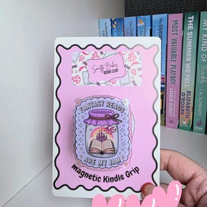 May include: A pink magnetic Kindle grip with a jar design. The jar contains a castle, a rainbow, and an open book with the text "FANTASY READS ARE MY JAM". The grip is on a pink card with the text "Smutty Babes Book Club".