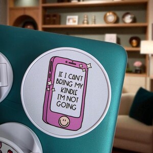 May include: A round white sticker with a pink smartphone graphic and the text "IF I CAN'T BRING MY KINDLE I'M NOT GOING." The smartphone has a smiling face at the bottom. The sticker is on a teal surface.