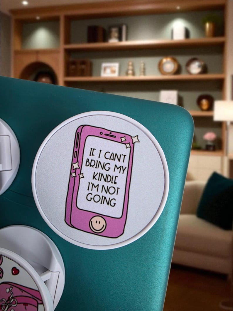 May include: A round white sticker with a pink smartphone graphic and the text "IF I CAN'T BRING MY KINDLE I'M NOT GOING." The smartphone has a smiling face at the bottom. The sticker is on a teal surface.