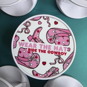 May include: White round phone grip with a pink cowboy-themed design. The design includes a pink cowboy hat, boots, and bows, with the text "WEAR THE HAT RIDE THE COWBOY".