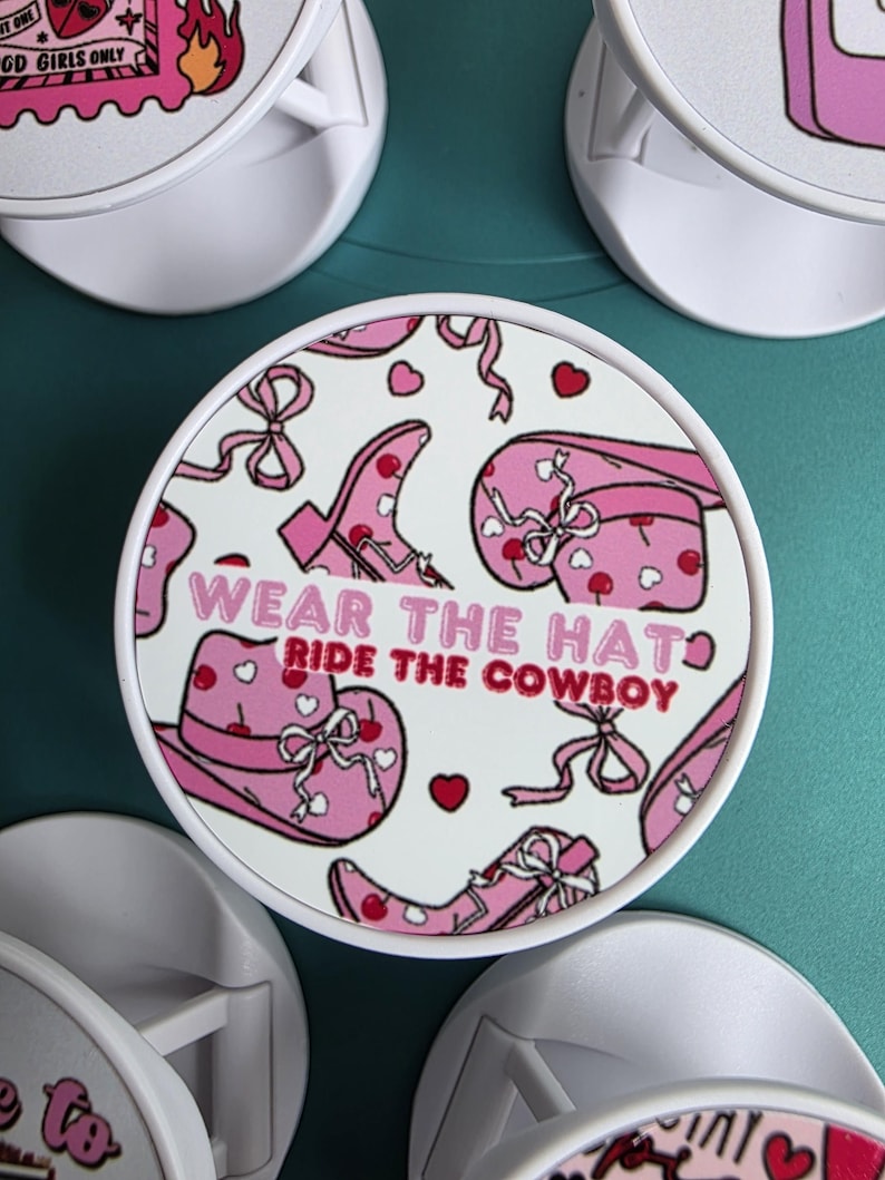 May include: White round phone grip with a pink cowboy-themed design. The design includes a pink cowboy hat, boots, and bows, with the text "WEAR THE HAT RIDE THE COWBOY".