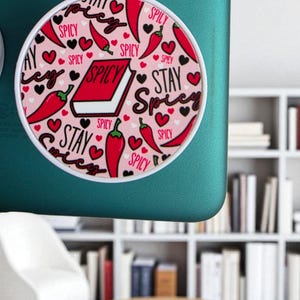 May include: A round, white-rimmed phone grip with a pink background. The design features red chili peppers, black hearts, and the words "SPICY" and "STAY SPICY" in a handwritten font. A red book with the word "SPICY" is also featured.