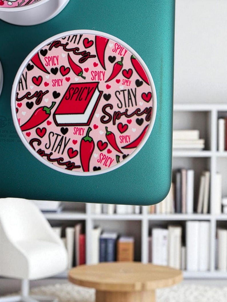 May include: A round, white-rimmed phone grip with a pink background. The design features red chili peppers, black hearts, and the words "SPICY" and "STAY SPICY" in a handwritten font. A red book with the word "SPICY" is also featured.