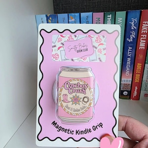 May include: A pink magnetic Kindle grip featuring a pink soda can design with the text "Cowboy Crush" and "Root & Rodeo Soda." The grip is on a pink background with the text "Magnetic Kindle Grip."