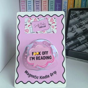 May include: A pink and white magnetic Kindle grip with the text "FOCK OFF I'M READING" in a speech bubble. The grip is attached to a pink card with the "Smutty Babes Book Club" logo. A Kindle and books are in the background.