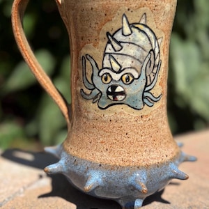 May include: A handmade ceramic mug with a tan speckled finish and a blue base with spike-like protrusions. The mug features a cartoon illustration of a blue creature with yellow eyes. The handle is a curved, tan color.