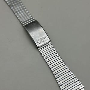 Puede incluir: Una correa de reloj extensible de color plateado con un cierre rectangular. El cierre está marcado con la palabra "Mido" en una fuente estilizada. La correa está hecha de pequeños eslabones metálicos articulados.