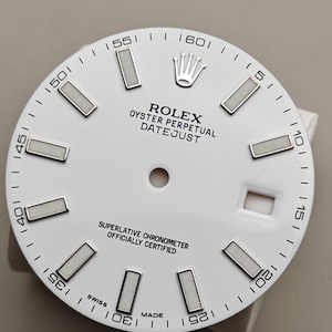 May include: A white Rolex Oyster Perpetual Datejust watch face with silver-colored rectangular hour markers and a date window. The watch face features the Rolex crown logo and the text "ROLEX OYSTER PERPETUAL DATEJUST". The words "SWISS MADE" are also visible.