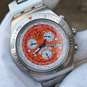 May include: A silver-toned Swatch chronograph watch with an orange dial and white sub-dials. The watch face displays the Swatch logo, time zones, and multiple hands. The watch has a silver band and a textured bezel.