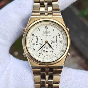 May include: A gold-toned Seiko Quartz Chronograph watch with a white face. The watch features multiple sub-dials, a tachymeter scale, and a gold link bracelet. The watch displays the day of the week and has a classic, vintage aesthetic.