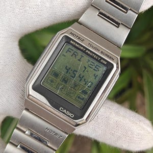 Puede incluir: Un reloj digital Casio Hotbiz vintage con una correa de metal plateado. La esfera del reloj muestra la hora y la fecha, con el texto "HOTBIZ TOUCH SCREEN MEMORY PROTECT 200" y "CASIO" visibles. El reloj se muestra sobre un fondo blanco.
