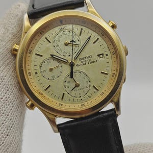 May include: A gold-toned Seiko Quartz World Timer watch with a black leather band. The watch face is cream-colored with multiple sub-dials and the text "SEIKO QUARTZ World Timer". The watch has a date display and is set against a neutral background.