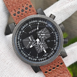 May include: A black Nixon watch with a brown leather strap. The watch face is black with white markings and hands. The strap has a perforated design and a buckle closure. The watch has a chronograph function.