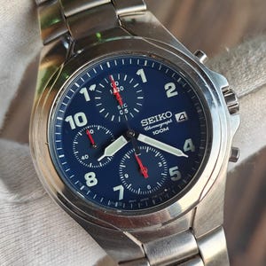 May include: A stainless steel Seiko Chronograph watch with a navy blue dial. The watch features white numerals, three sub-dials, and red hands. The watch band is made of stainless steel. The watch face reads "SEIKO Chronograph 100M".