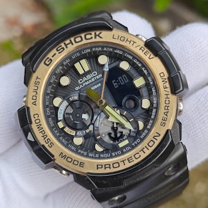 May include: A black and gold G-Shock Casio Gulfmaster watch. The watch face displays the time as 6:00, with multiple dials and text including "COMPASS", "MODE", and "PROTECTION". The watch has a black band and a gold bezel.